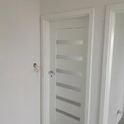 Białe, nowoczesne drzwi wewnętrzne z poziomymi przeszkleniami, zamontowane w surowym wnętrzu, widoczne niedokończone ściany i wystające przewody elektryczne.