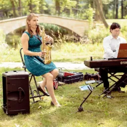 Zespół muzyczny grający na saksofonie i keyboardzie na tle parku z kamiennym mostkiem.