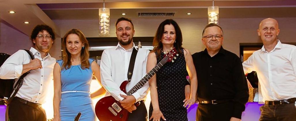 Sześcioosobowy skład muzyczny pozuje z instrumentami, w tym z czerwoną gitarą elektryczną, na tle sali z dekoracyjnym oświetleniem.