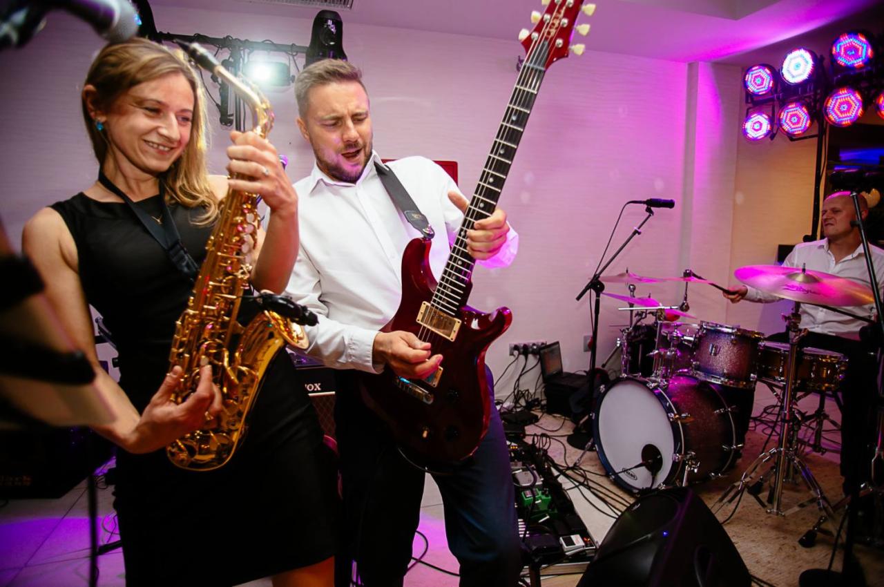 Saksofonistka w czarnej sukience i gitarzysta grający na czerwonej gitarze elektrycznej, z perkusistą w tle, podczas występu na żywo z oświetleniem scenicznym.
