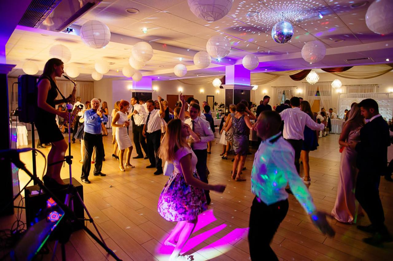 Saksofonistka grająca na scenie podczas imprezy, tańczący goście w tle, dekoracyjne lampiony i kula dyskotekowa.