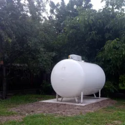 Biały, poziomy zbiornik na gaz LPG ustawiony na betonowej podstawie w ogrodzie, otoczony drzewami.
