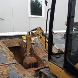 Żółta mini koparka CAT pogłębia wykop w ziemi przy budynku z szarej blachy trapezowej i białym oknie.