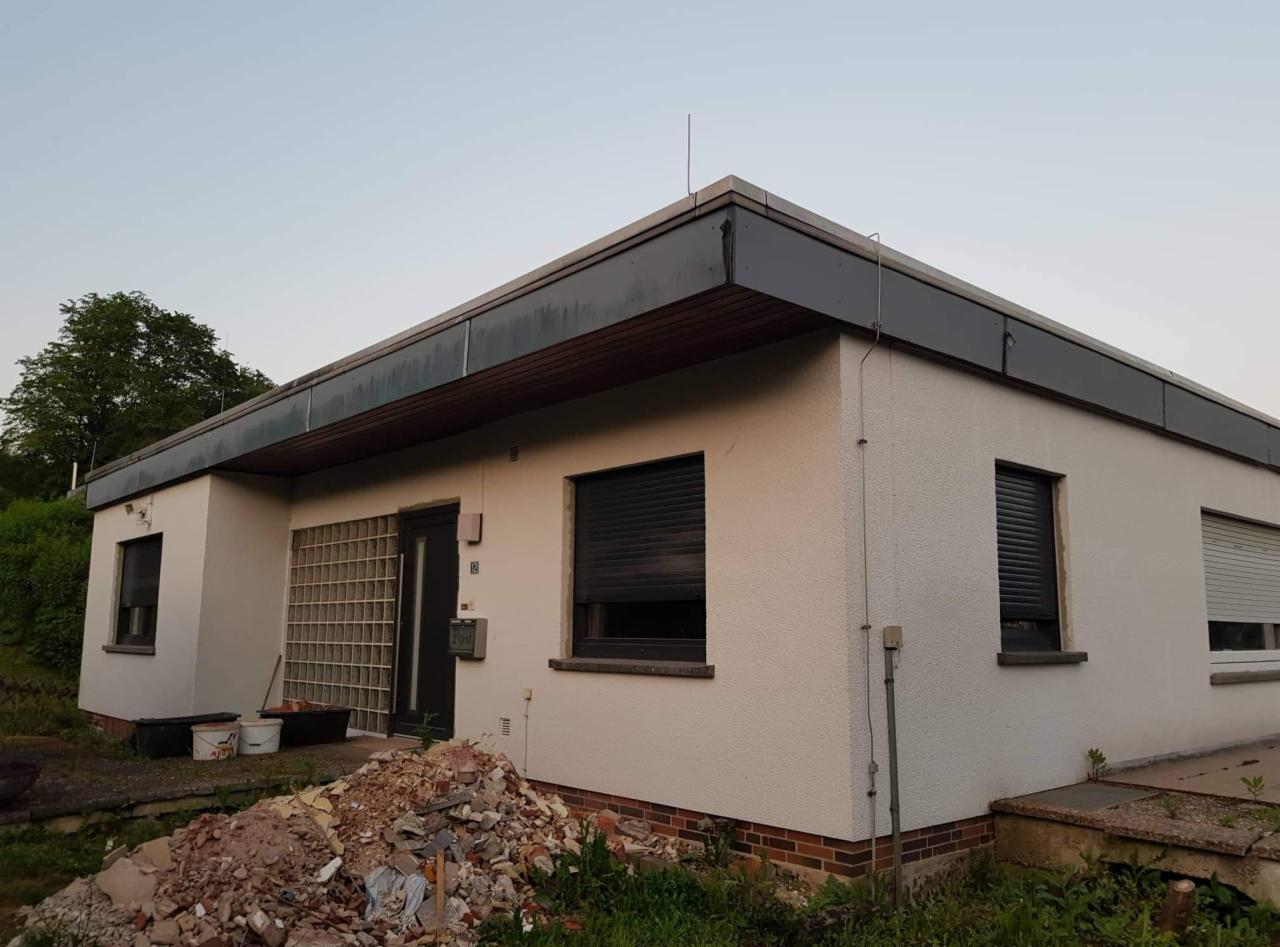 Dom jednorodzinny z płaskim dachem, widoczne ślady prac remontowych i gruz przed budynkiem, częściowo zasłonięte okna roletami zewnętrznymi.