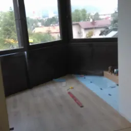 Układanie paneli na balkonie z widokiem na miasto. Widoczne narzędzia i podkład izolacyjny. Częściowo ukończona podłoga z paneli.