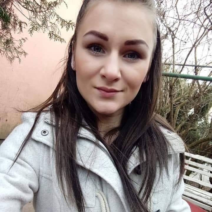 Portret kobiety w jasnym płaszczu na tle ściany i drzew. Uśmiechnięta twarz, długie włosy, naturalny makijaż. Zdjęcie z zewnątrz, w tle gałęzie i ławka.