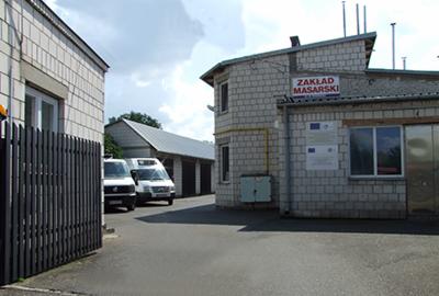 Budynek Zakładu Masarskiego w Garwolinie z zaparkowanymi samochodami dostawczymi, widoczne tablice informacyjne i charakterystyczna biała cegła.