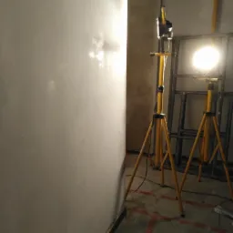 Świeżo wykończona ściana oświetlona dwoma lampami na żółtych statywach, widoczne ślady po szpachlowaniu i przygotowaniu do dalszych prac wykończeniowych.