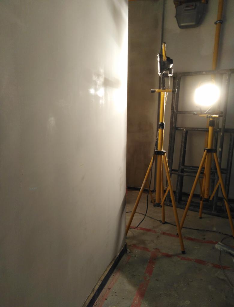 Świeżo wykończona ściana oświetlona dwoma lampami na żółtych statywach, widoczne ślady po szpachlowaniu i przygotowaniu do dalszych prac wykończeniowych.