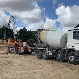 Ciężarówka z betonomieszarką Eurobud i pompa do betonu na placu budowy, w tle drzewa i błękitne niebo z chmurami.