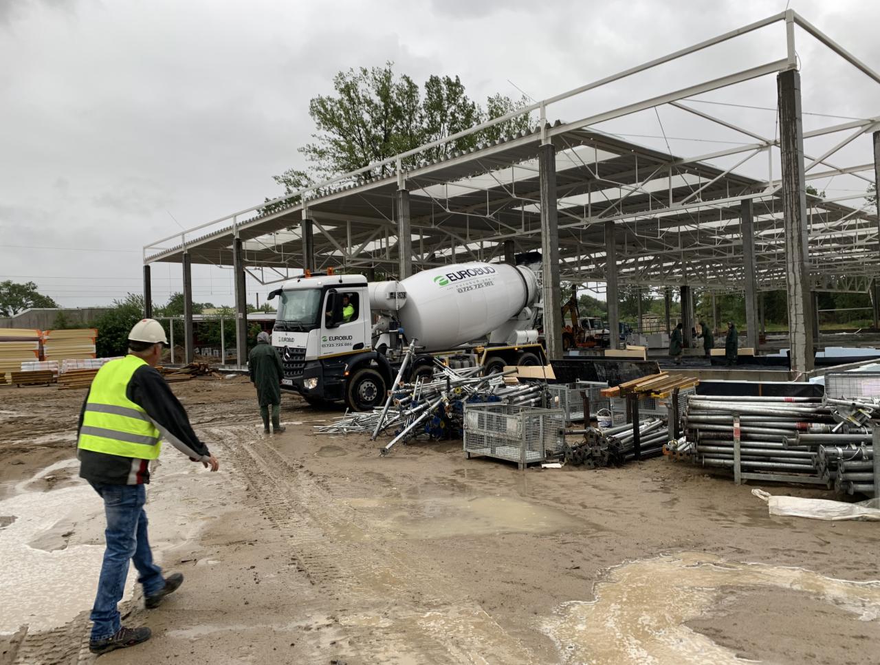 Budowa hali stalowej z betonowymi fundamentami, widoczny mikser do betonu i pracownicy w odblaskowych kamizelkach na błotnistym terenie.