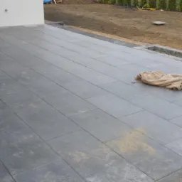 Układanie szarej kostki betonowej na tarasie, widoczne niedociągnięcia w postaci piasku i tkaniny budowlanej.