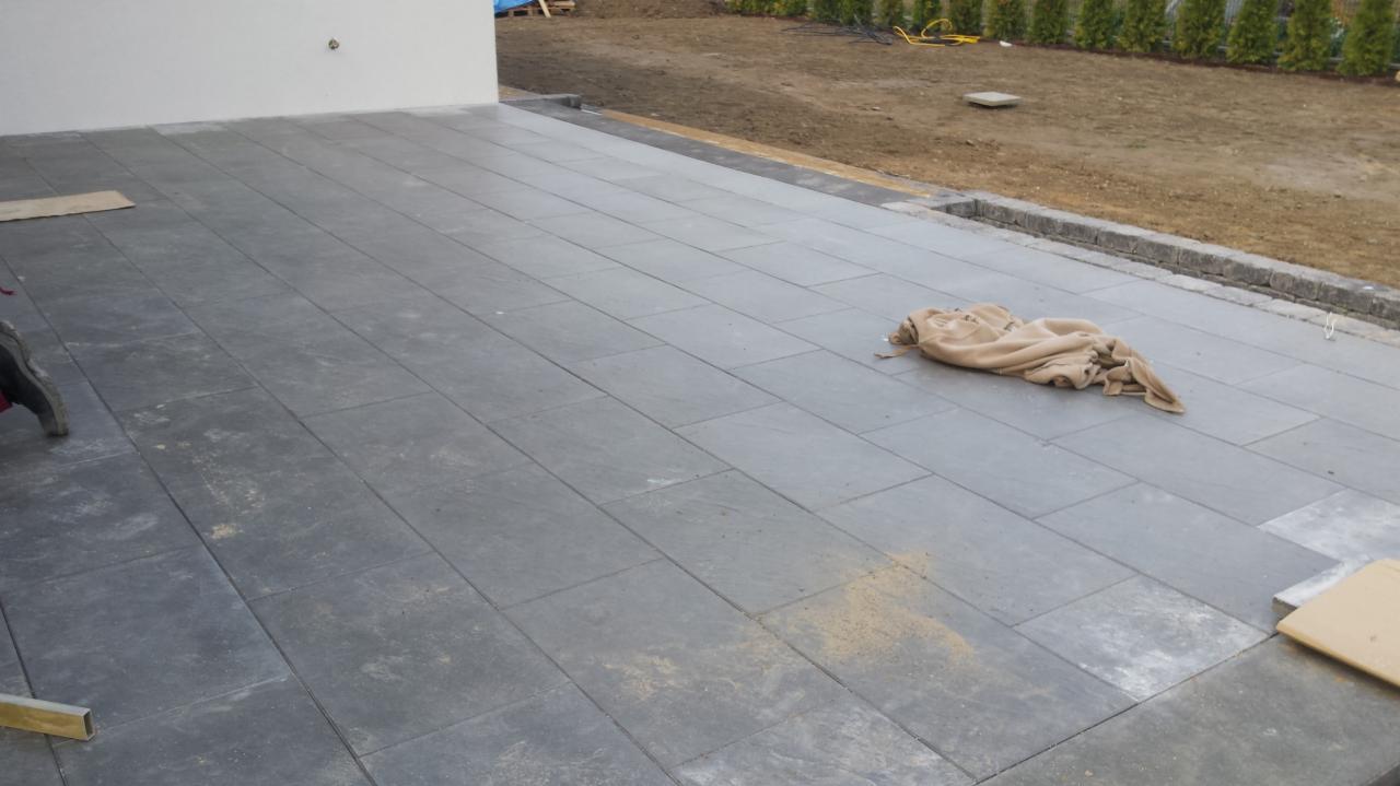 Układanie szarej kostki betonowej na tarasie, widoczne niedociągnięcia w postaci piasku i tkaniny budowlanej.