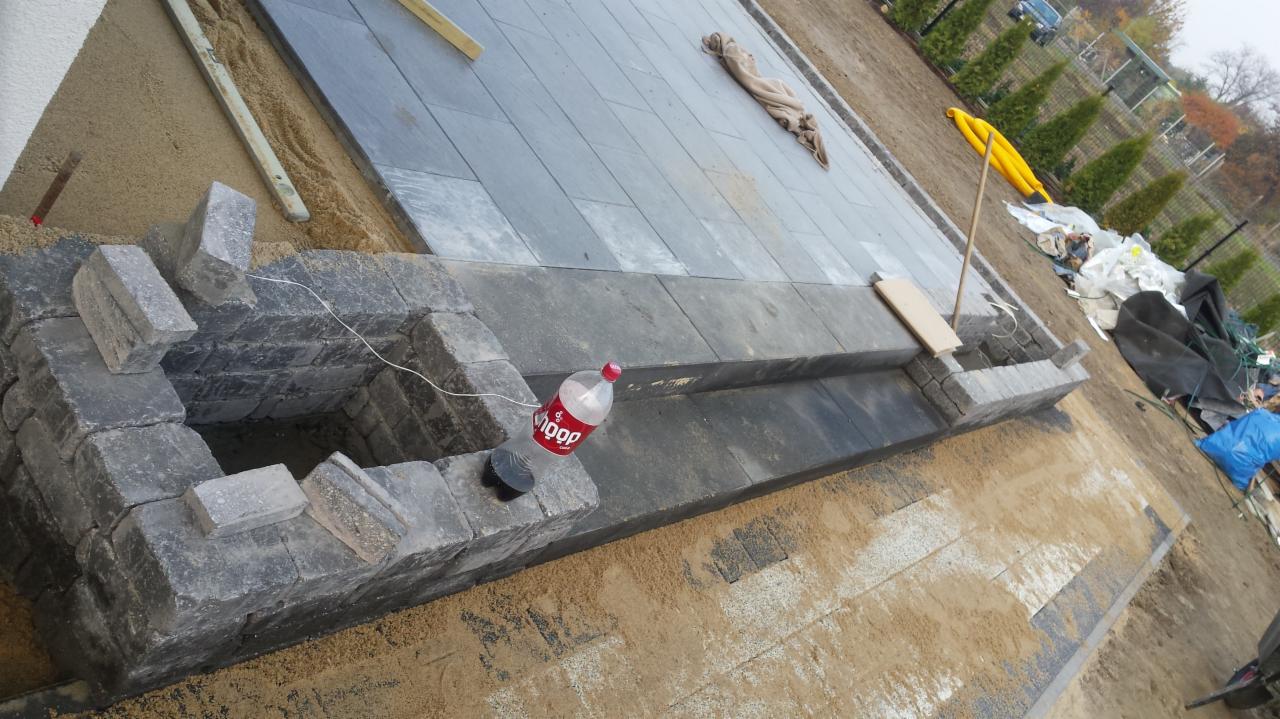 Budowa nawierzchni z szarych płyt betonowych i obrzeża z kamiennych bloczków, widoczna butelka po napoju i piasek zasypujący szczeliny.