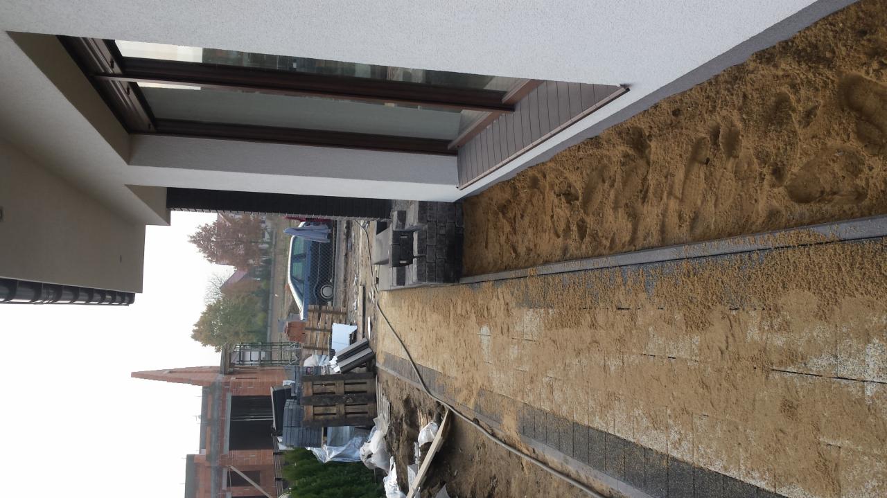 Przygotowanie podłoża pod nawierzchnię z kostki brukowej, widoczne warstwy piasku i podsypki, w tle budowa domu.