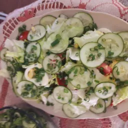 Sałatka warzywna z jajkiem, pomidorem, ogórkiem i szczypiorkiem, podana na owalnym talerzu na tle wzorzystego obrusa.