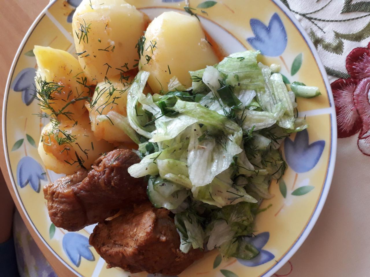 Talerz z obiadem: ziemniaki z koperkiem, sałatka z ogórkiem i mięsne kotlety na talerzu w kwiatki, widok z góry.