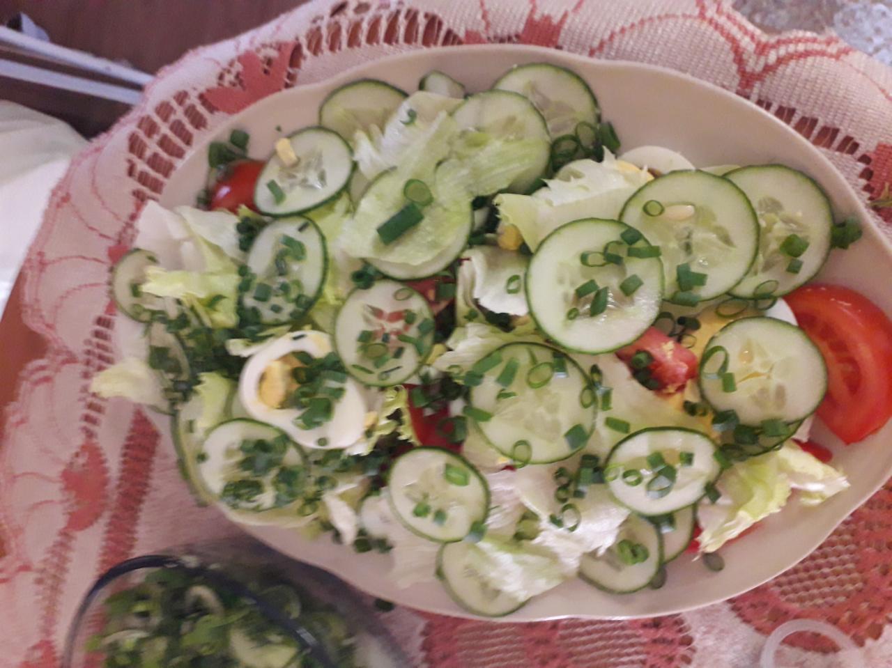 Sałatka warzywna z jajkiem, pomidorem, ogórkiem i szczypiorkiem, podana na owalnym talerzu na tle wzorzystego obrusa.