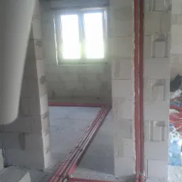 Czerwone rury instalacyjne ułożone wzdłuż ścian i podłogi w surowym wnętrzu budynku w Rabce-Zdroju, widoczne przez otwór drzwiowy, z oknem w tle.