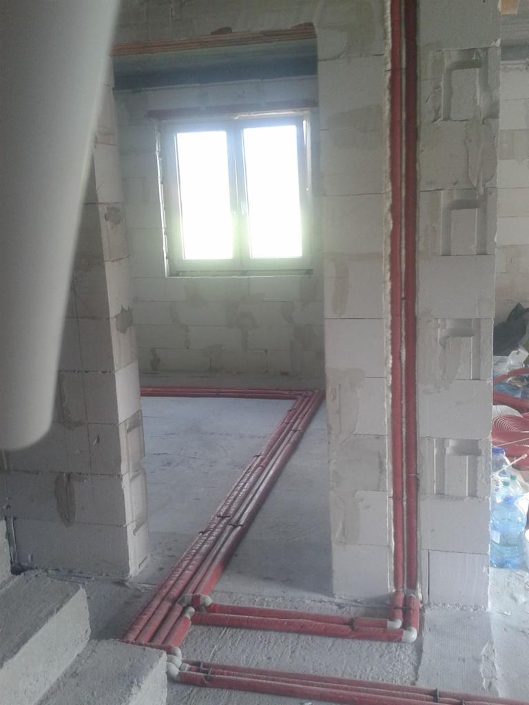 Czerwone rury instalacyjne ułożone wzdłuż ścian i podłogi w surowym wnętrzu budynku w Rabce-Zdroju, widoczne przez otwór drzwiowy, z oknem w tle.