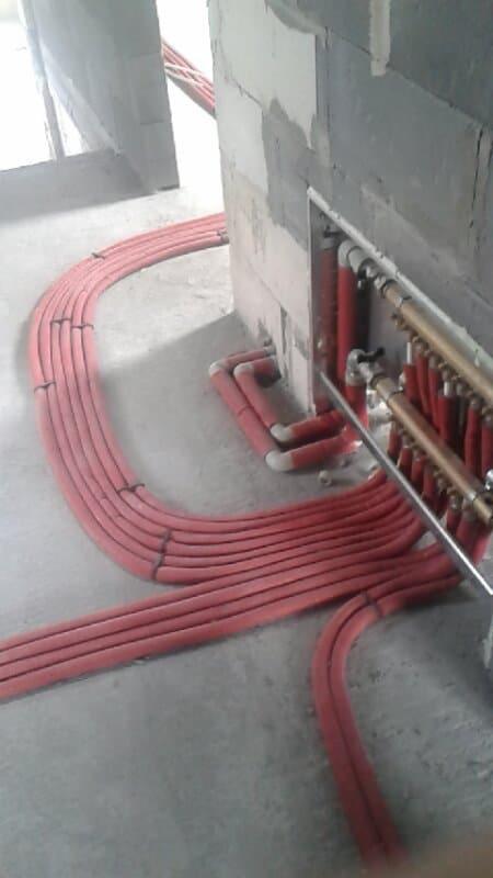Rozprowadzenie czerwonych rur instalacji ogrzewania podłogowego w surowym stanie, widoczne złącza i rozdzielacz na ścianie.