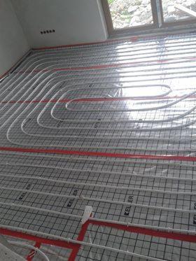 Rozłożona instalacja ogrzewania podłogowego z białych rur na siatce, widoczne czerwone elementy mocujące, surowy stan wykończenia pomieszczenia.