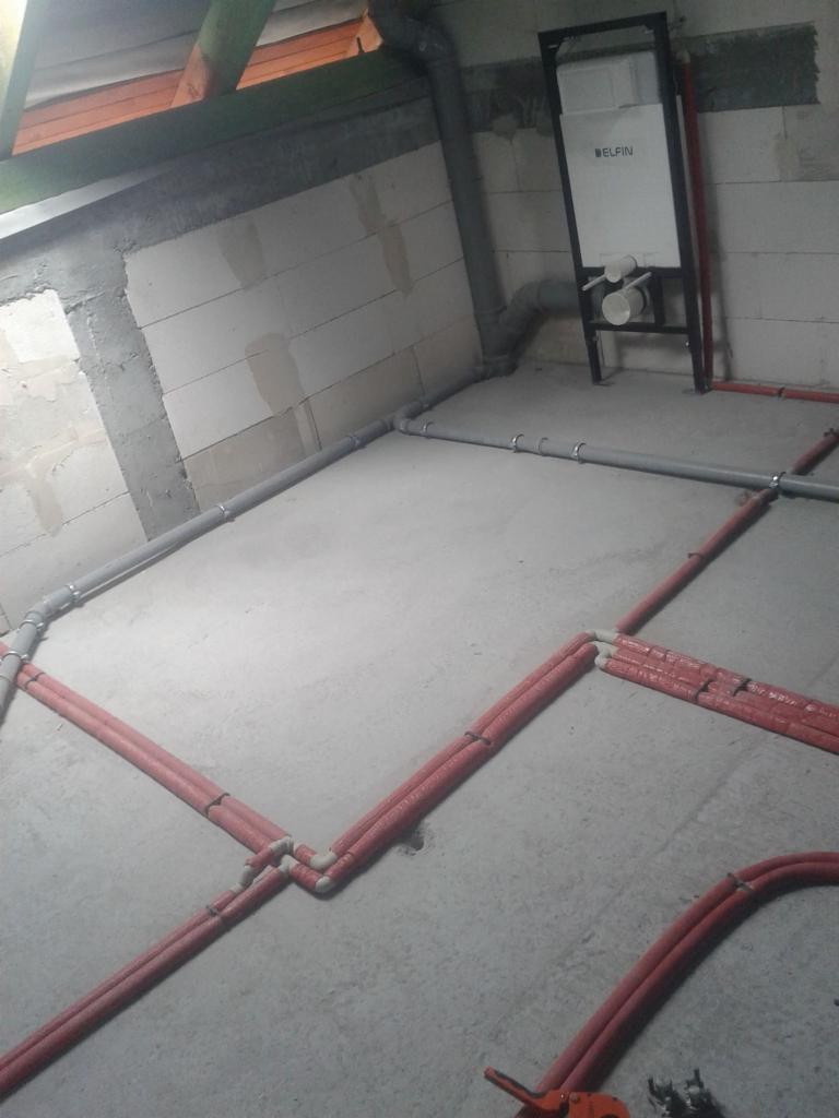 Surowy stan instalacji wodnej i kanalizacyjnej w nowym budynku: czerwone rury w izolacji na podłodze, szare rury kanalizacyjne przy ścianie z bloczków betonowych, stelaż podtynkowy WC z widocznym...