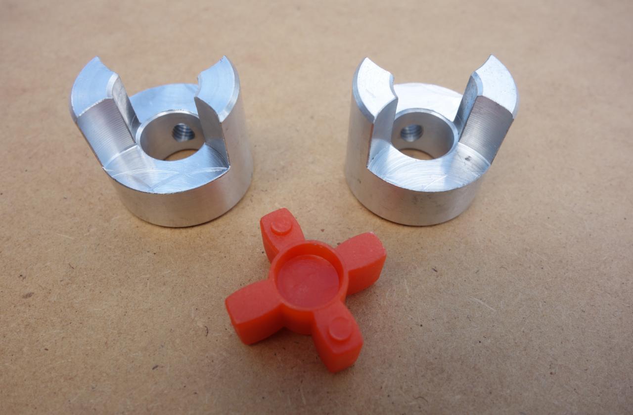 Dwa aluminiowe półprodukty z wyfrezowanymi rowkami i gwintowanym otworem oraz czerwony, gumowy element sprzęgła na brązowym tle.