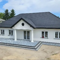 Nowoczesny dom jednorodzinny z białymi ścianami i ciemną dachówką, z tarasem wyłożonym szarą kostką brukową i trzema schodami wejściowymi, widok z lotu ptaka na częściowo zagospodarowany teren.