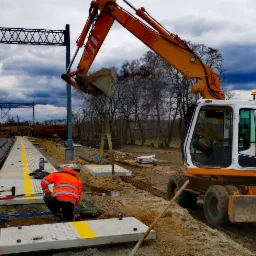 Prace ziemne przy budowie peronu kolejowego: robotnicy w odblaskowych ubraniach, koparka Liebherr podnosi betonowy element, w tle torowisko i pochmurne niebo.