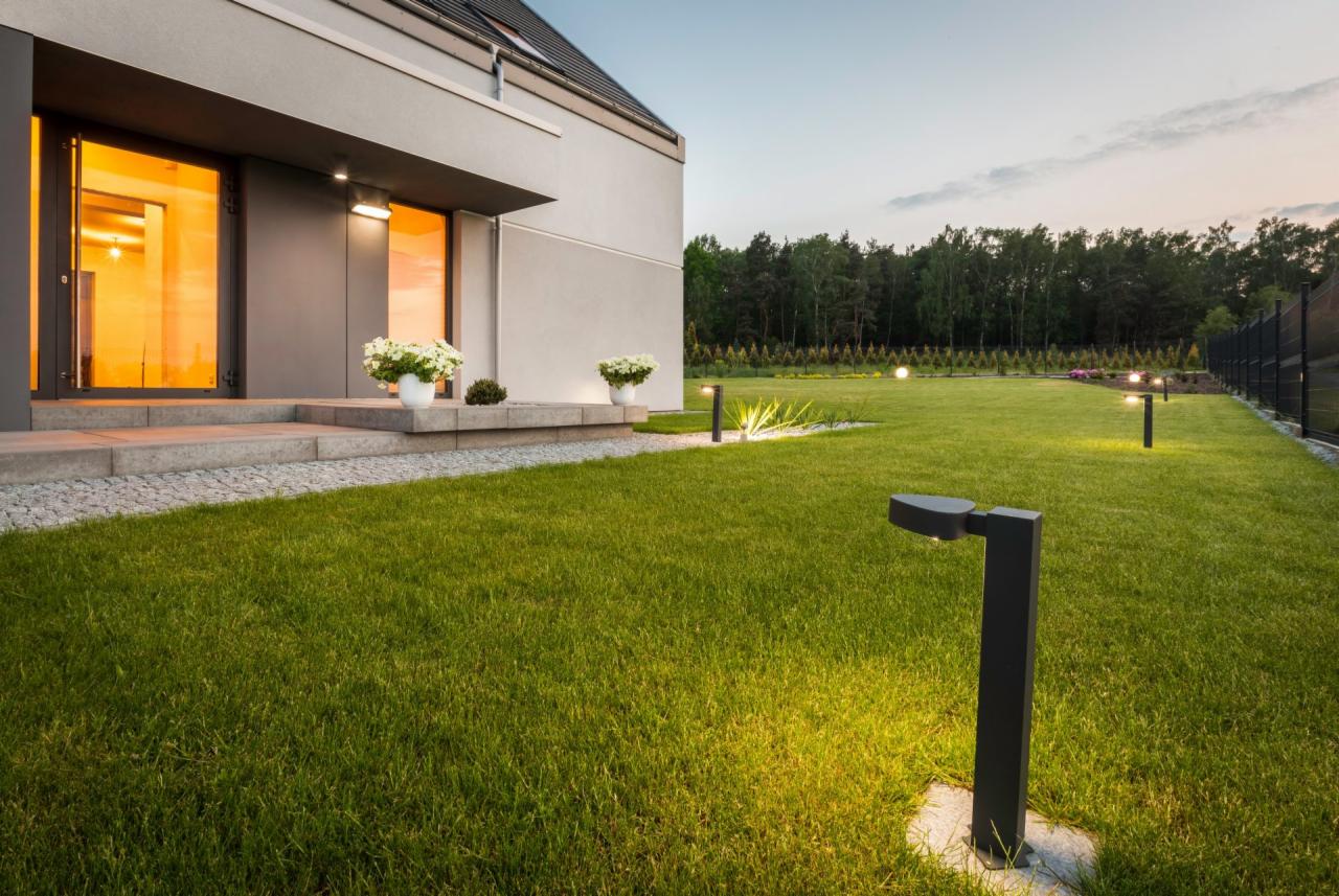 Nowoczesny dom z oświetlonym ogrodem, lampy ogrodowe wzdłuż trawnika, kwiaty w donicach przy wejściu, wieczorny krajobraz.