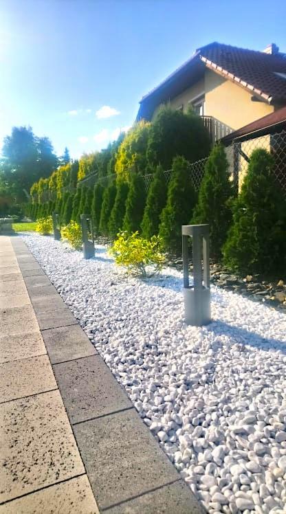 Ścieżka z kostki brukowej obok białych kamieni i szpaleru tuj z oświetleniem ogrodowym, widok na dom w tle.