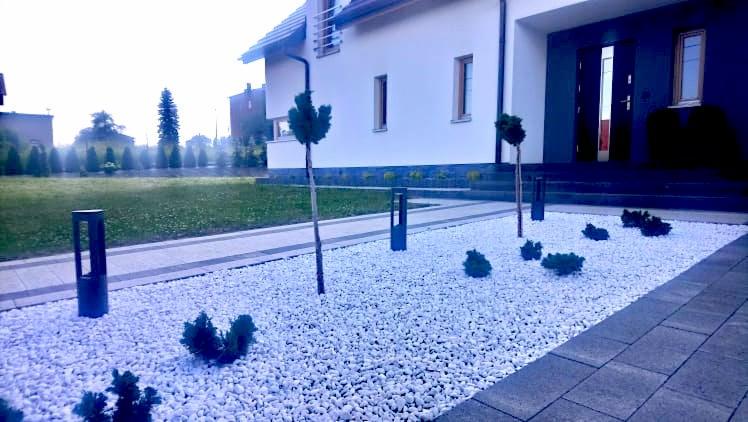 Nowoczesny ogród z białym żwirem, niskimi krzewami, ścieżką z płyt i minimalistycznymi lampami ogrodowymi na tle domu z ciemnymi oknami i drzwiami.