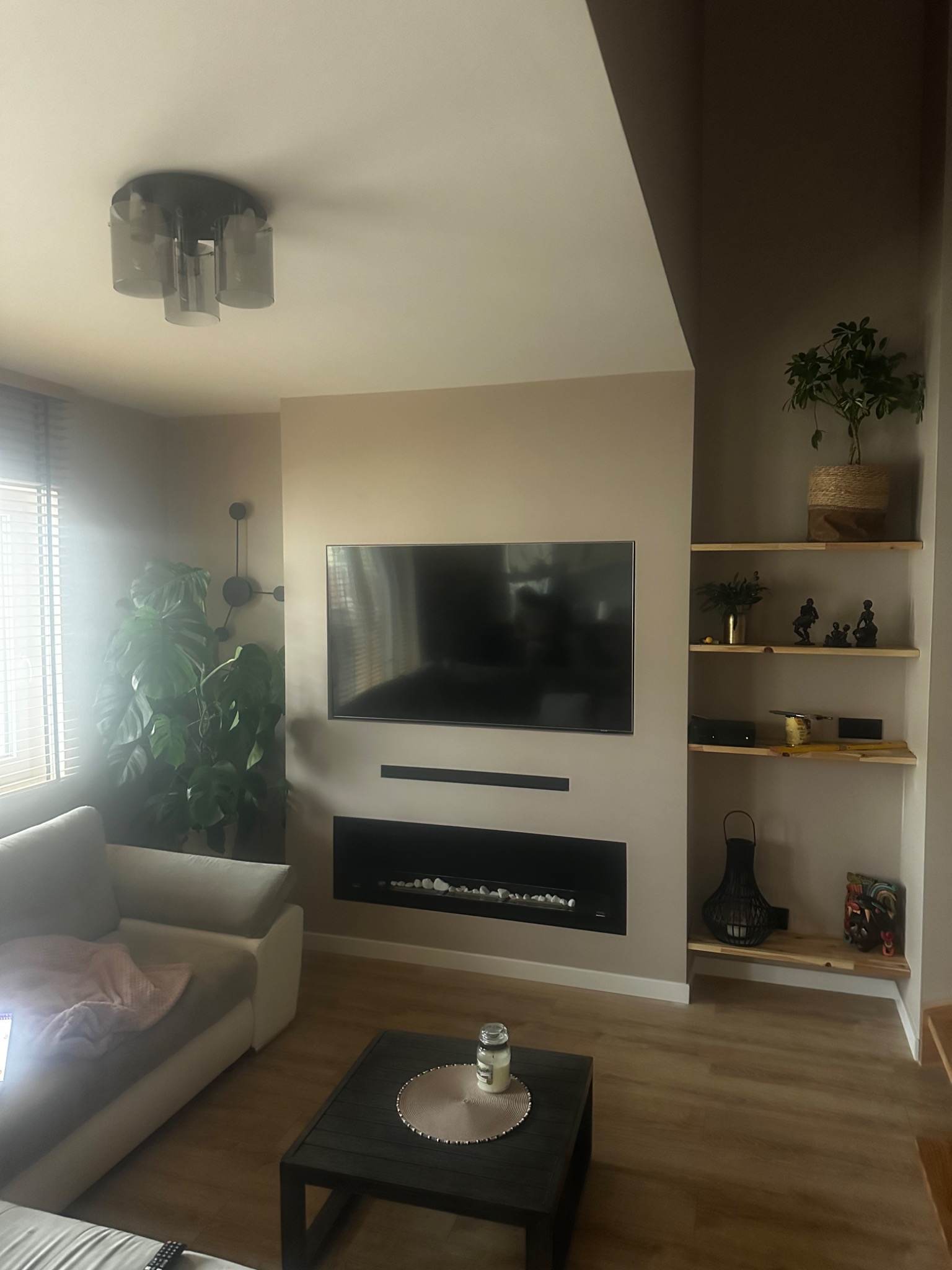 Nowoczesny salon z beżowymi ścianami, podwieszonym telewizorem nad elektrycznym kominkiem z białymi kamieniami, drewnianą podłogą i półkami z dekoracjami, oświetlony przez okno z żaluzjami i lampę...