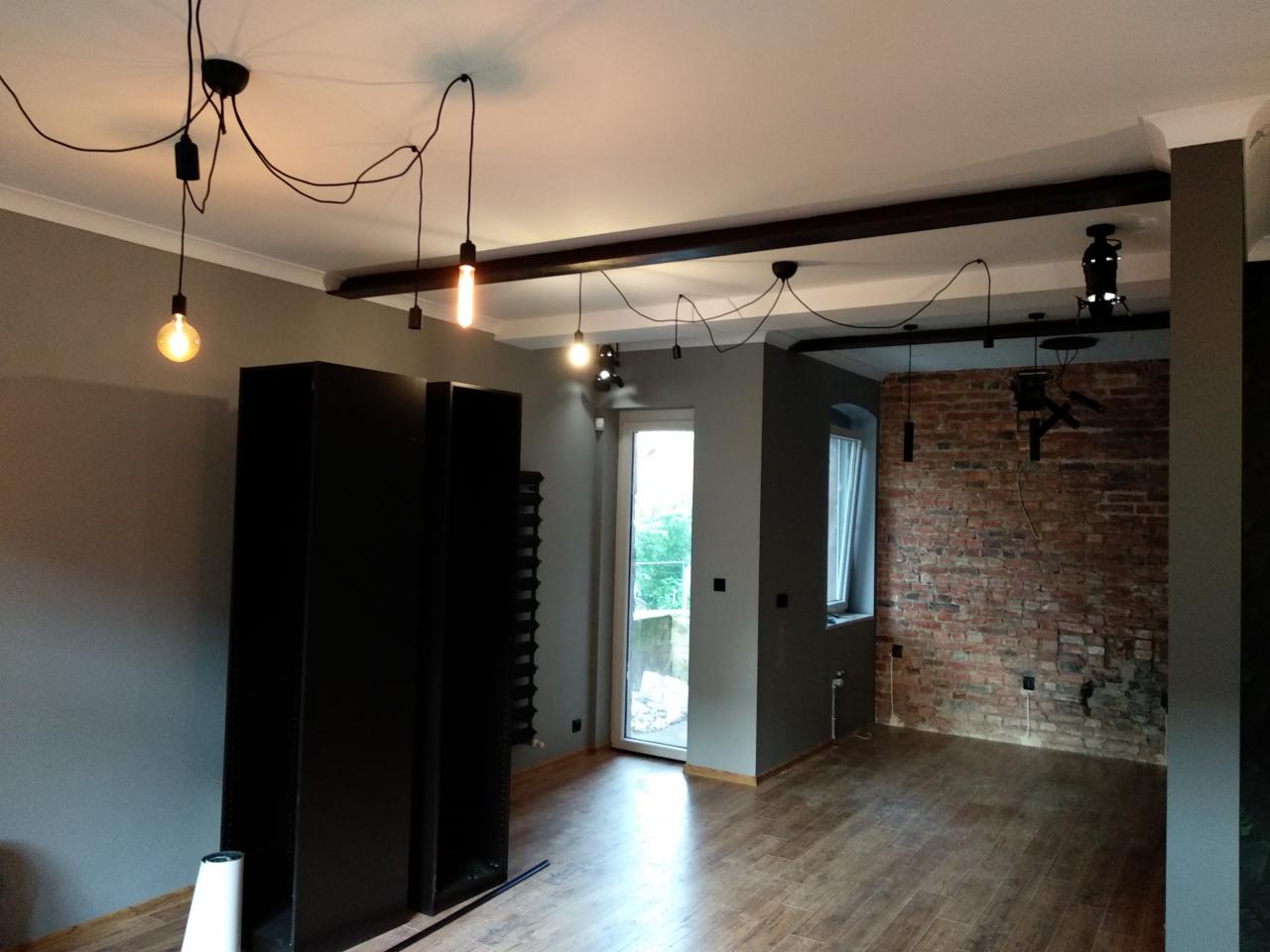 Pokój w trakcie urządzania: widoczna ceglana ściana, drewniana podłoga, industrialne oświetlenie z żarówkami Edisona na kablach, czarne regały i szare ściany.