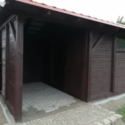 Ciemnobrązowa konstrukcja drewniana z czerwonym dachem, częściowo otwarta, z podłogą wyłożoną jasnoszarą kostką brukową, widoczna boczna ściana z małym oknem.