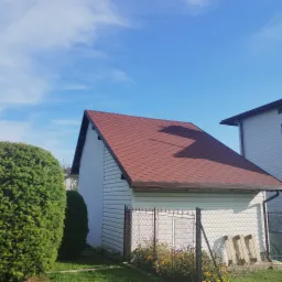 Dom z nowym, czerwonym dachem pokrytym gontem bitumicznym, widok zza metalowego ogrodzenia porośniętego żółtymi kwiatami.