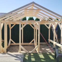 Kamil Pieczka - Konstrukcja z jasnego drewna, prawdopodobnie altany, w trakcie budowy. Widoczny szkielet z belek, częściowo zmontowany, z zielonymi krzewami w tle i niebieskim niebem. Na drabinie stoi osoba...