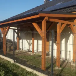 Drewniana konstrukcja zadaszenia tarasu przy domu jednorodzinnym, widoczna od strony ogrodu, z panelami słonecznymi na dachu budynku.