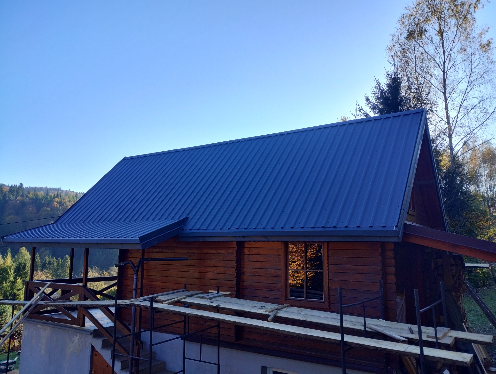 Nowy, ciemnoniebieski dach z blachy trapezowej na drewnianym domu w stylu góralskim, widoczne rusztowania i las w tle.