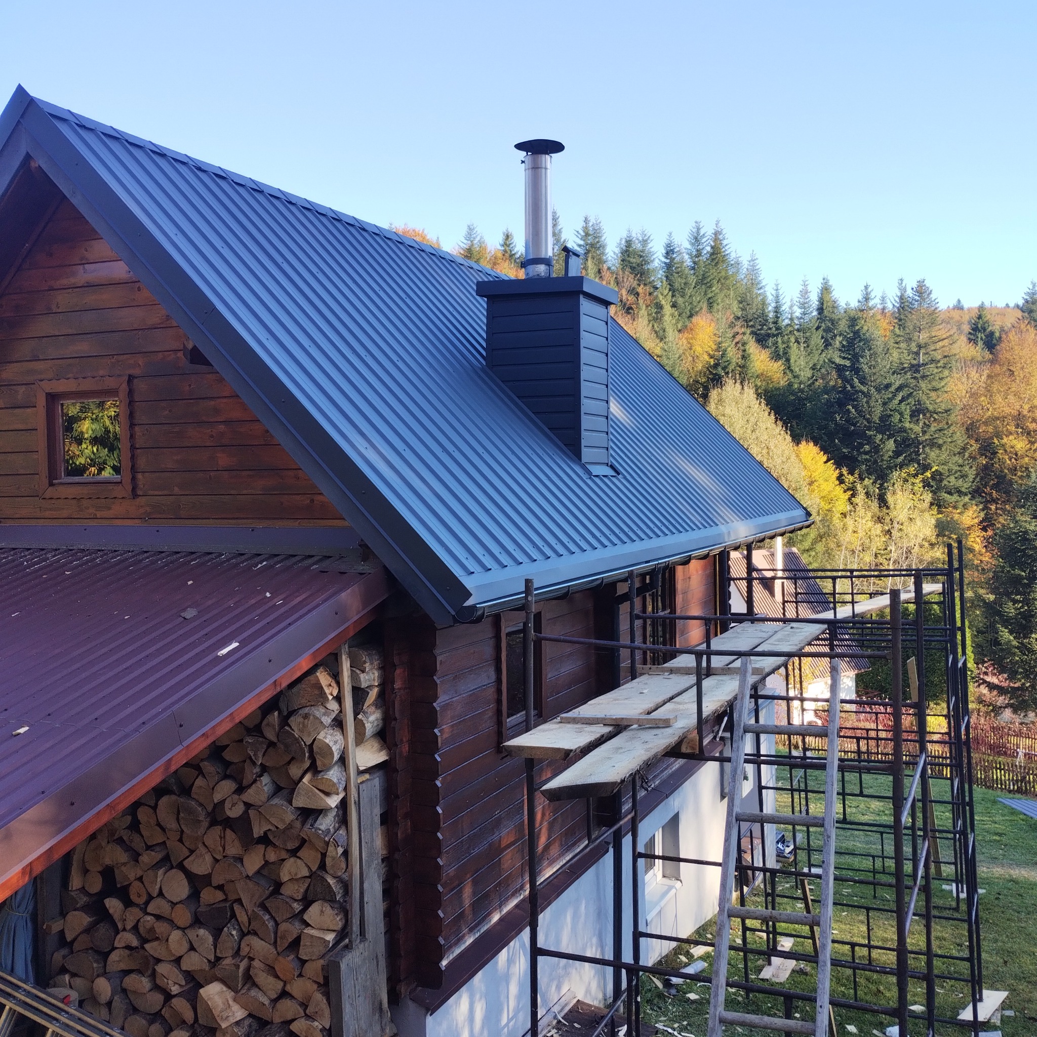 Dom z bali z nowym, ciemnoszarym dachem z blachy trapezowej i kominem, widok z rusztowania, w tle las z jesiennymi kolorami.