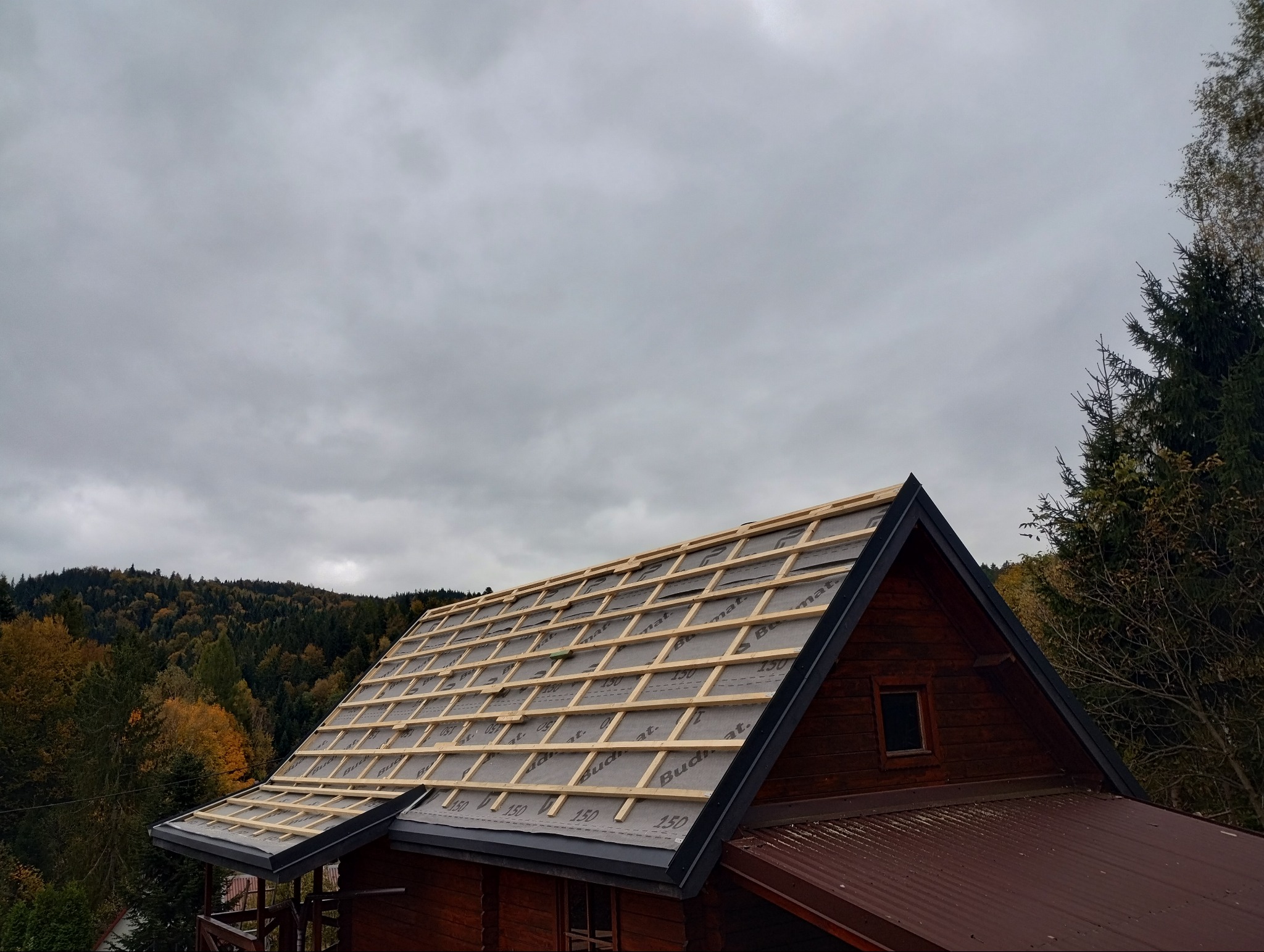 Wymiana poszycia dachu na drewnianym domu w górach, widoczne łaty i membrana dachowa przed położeniem dachówki, pochmurne niebo w tle.