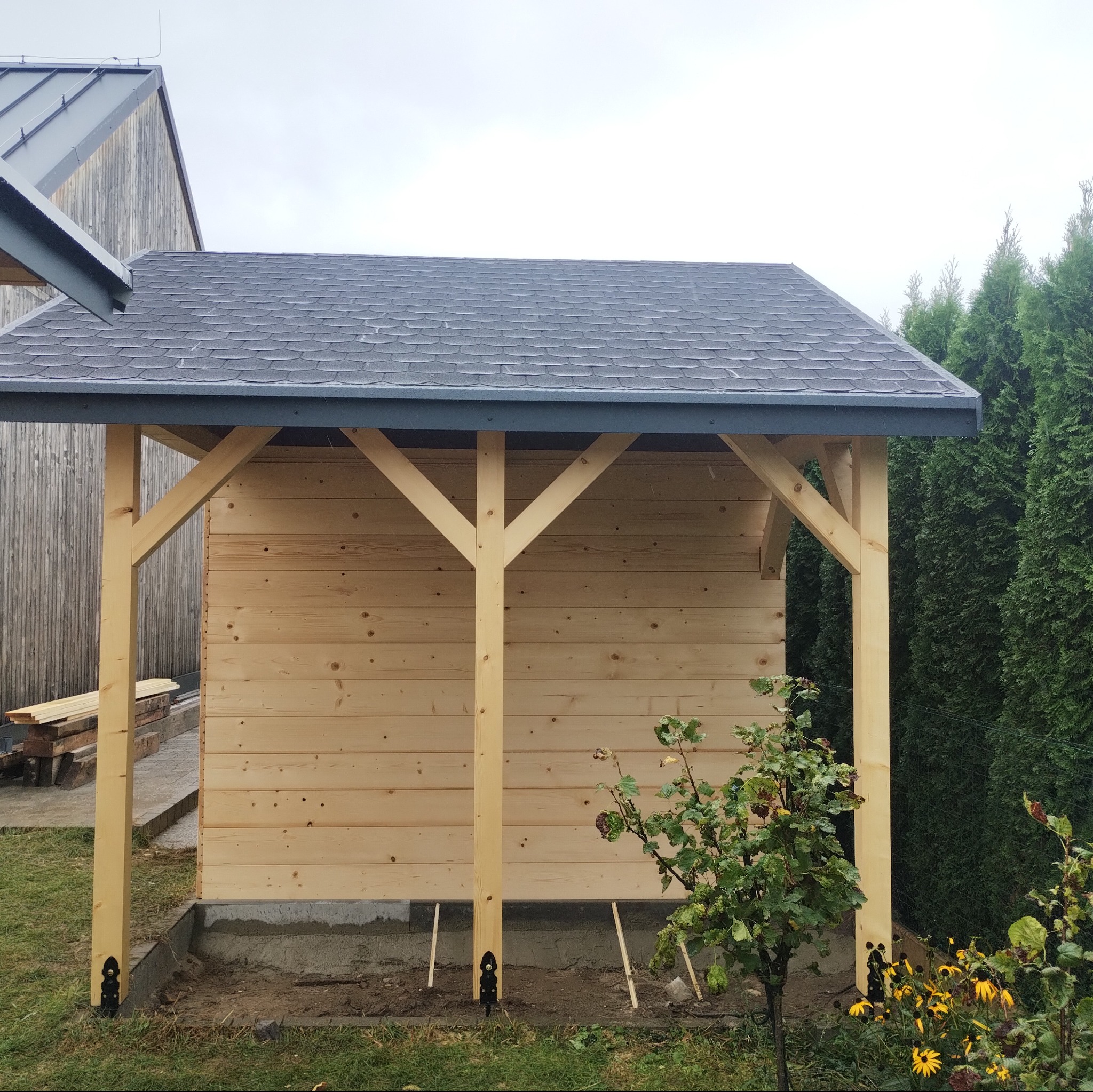Drewniana konstrukcja z szarym dachem z gontu bitumicznego, częściowo obita deskami, z widocznymi elementami mocującymi do podłoża, na tle zielonych tuj i drewnianego budynku.
