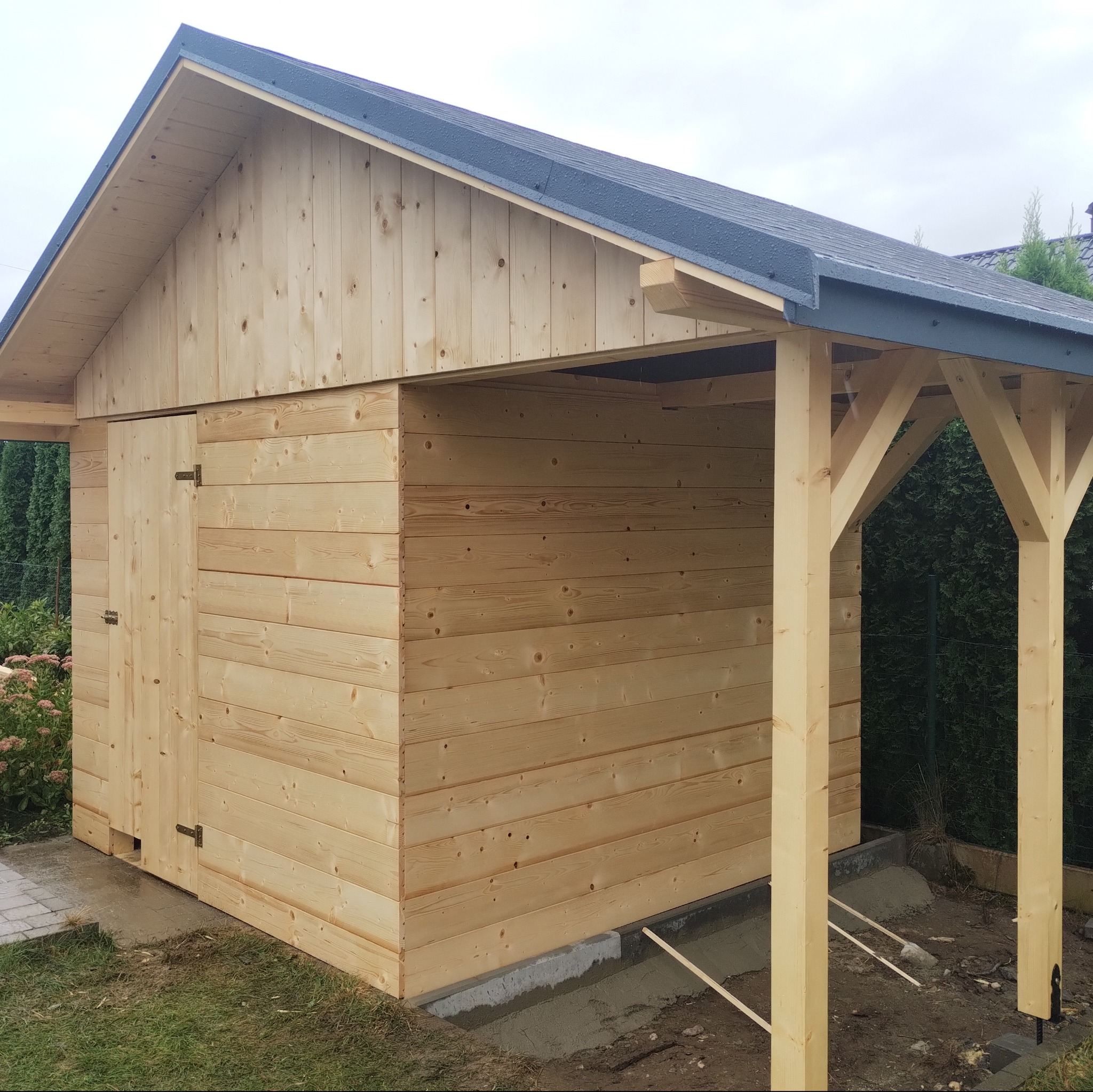 Nowa, surowa altana z drewna sosnowego z drzwiami i zadaszeniem, osadzona na betonowej wylewce, z widocznymi podporami i elementami konstrukcyjnymi, na tle zieleni.