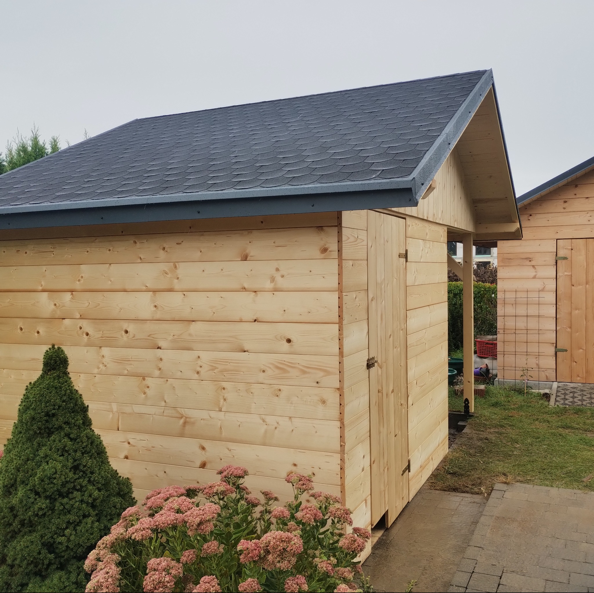 Dwie drewniane konstrukcje z bitumicznym pokryciem dachowym, otoczone zielenią, z widocznymi drzwiami i roślinnością w ogrodzie.