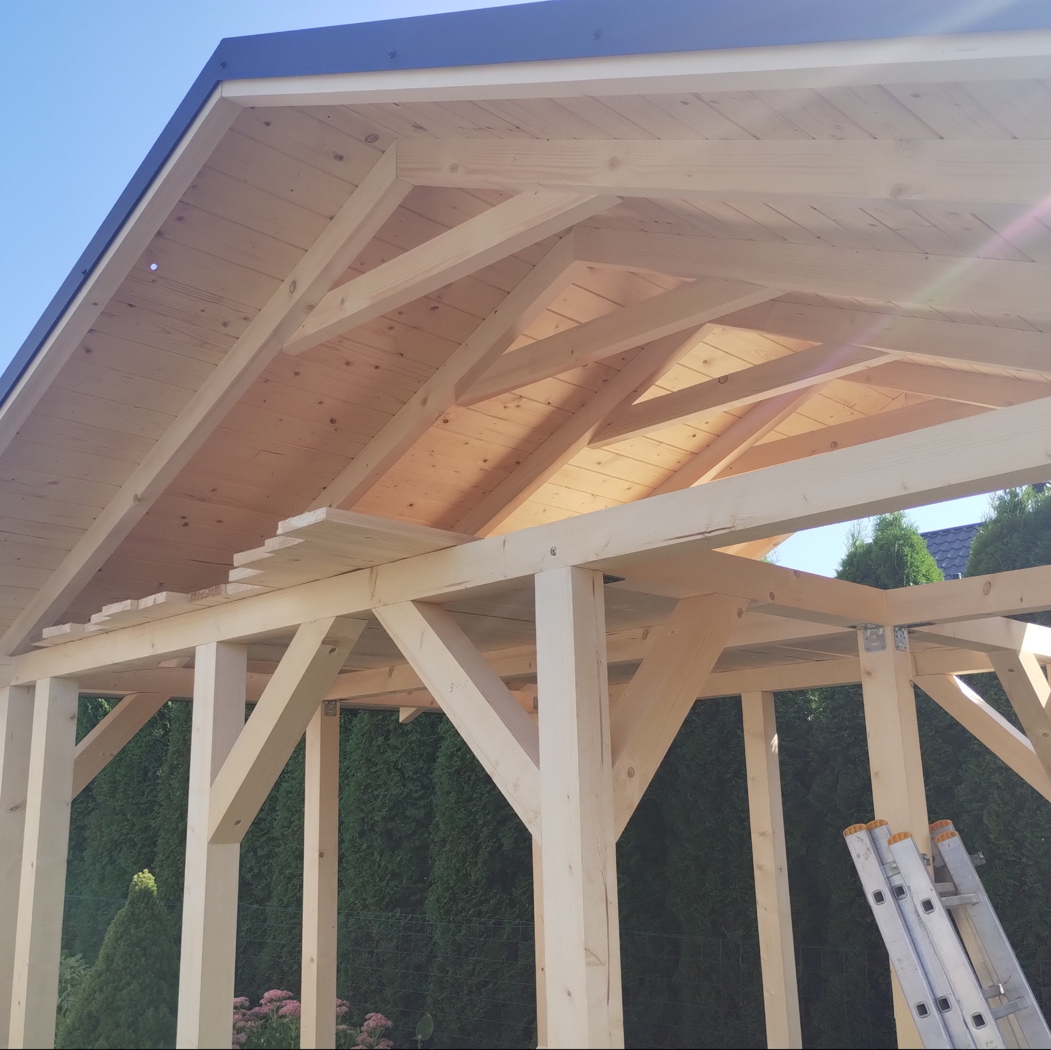Konstrukcja z jasnego drewna z elementami dachu w trakcie budowy, widoczna drabina oparta o konstrukcję, zieleń w tle.