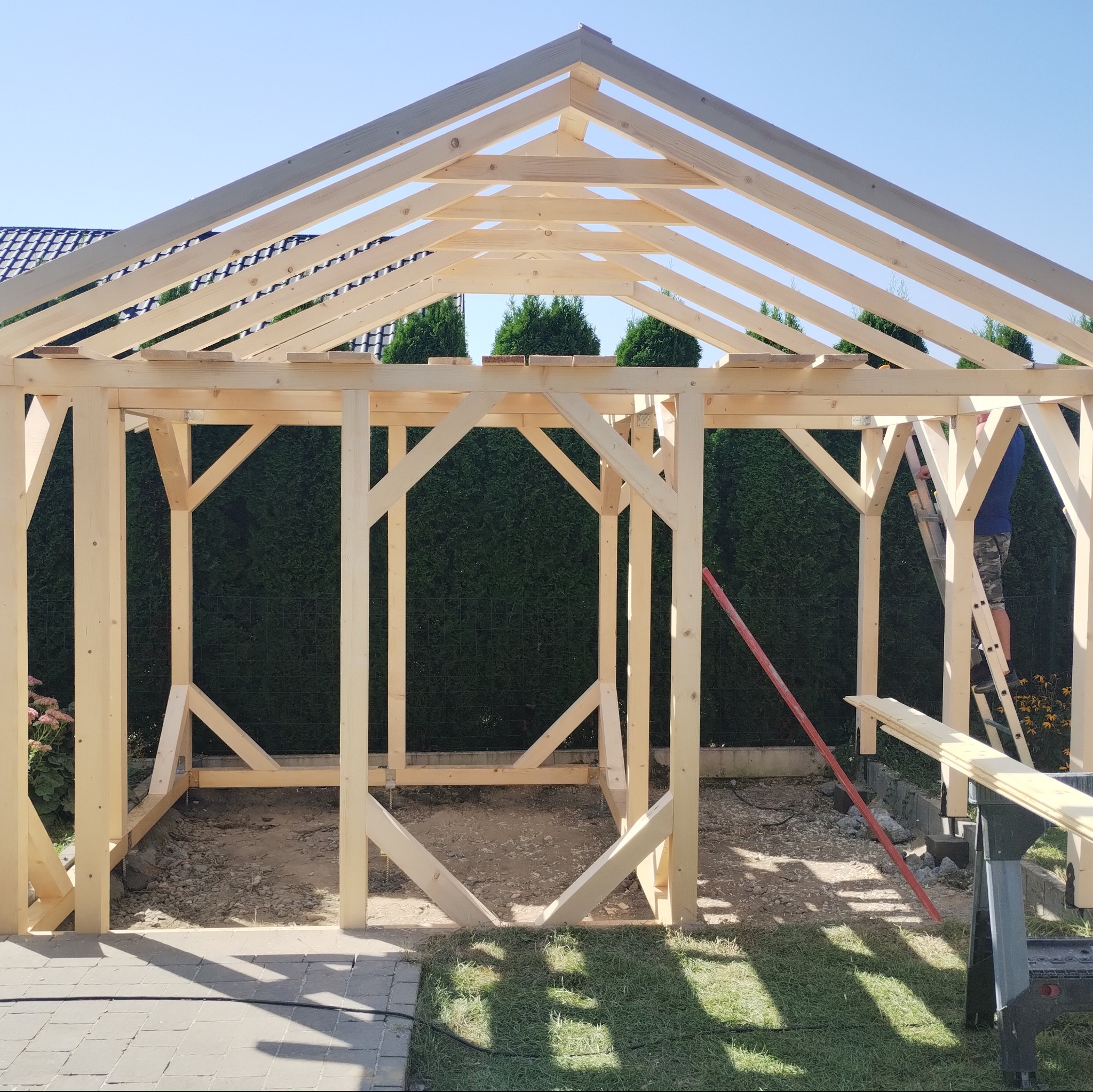 Konstrukcja z jasnego drewna, prawdopodobnie altany, w trakcie budowy. Widoczny szkielet z belek, częściowo zmontowany, z zielonymi krzewami w tle i niebieskim niebem. Na drabinie stoi osoba...