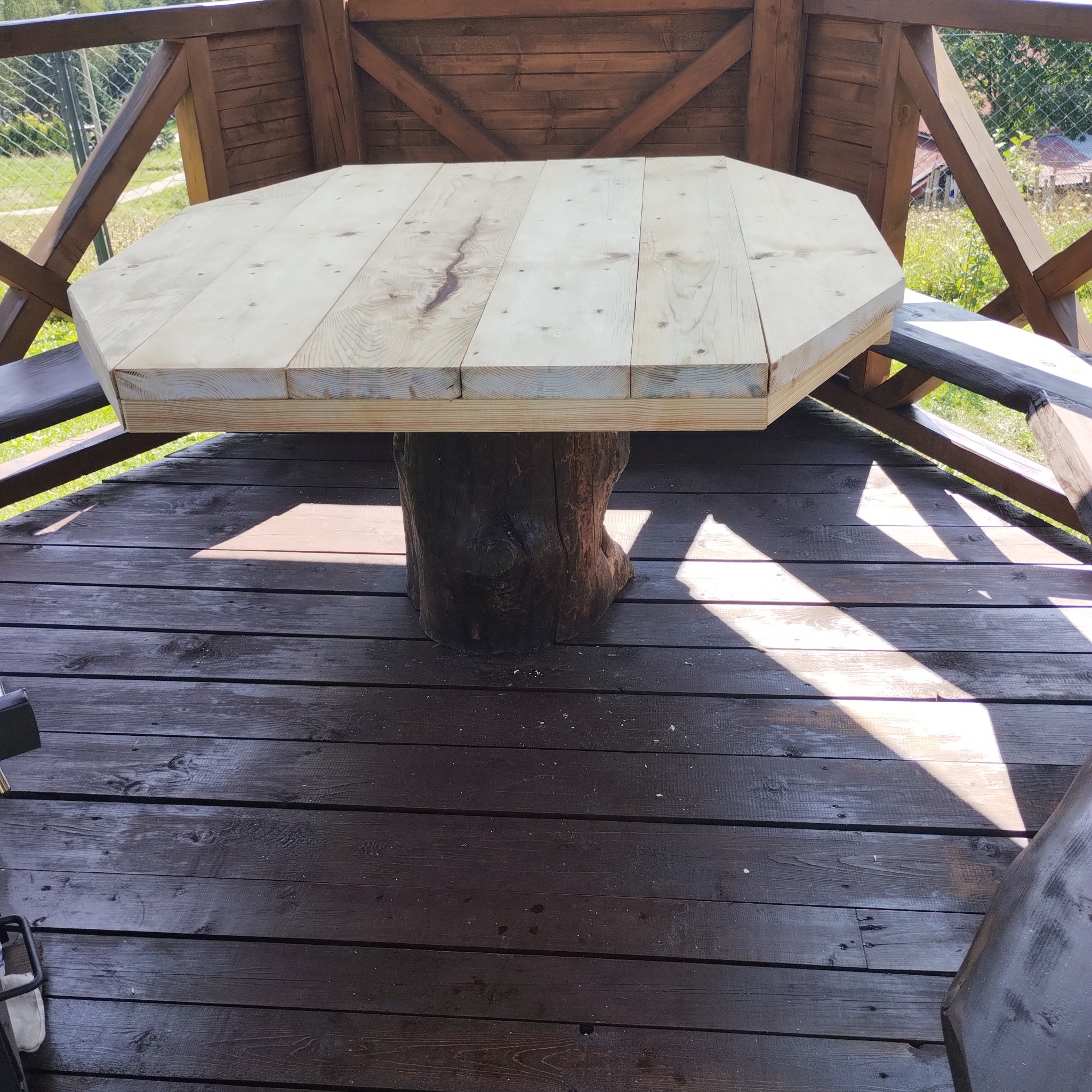 Ośmiokątny stół z jasnego drewna, osadzony na pniu drzewa wewnątrz drewnianej altany z widokiem na zewnątrz.