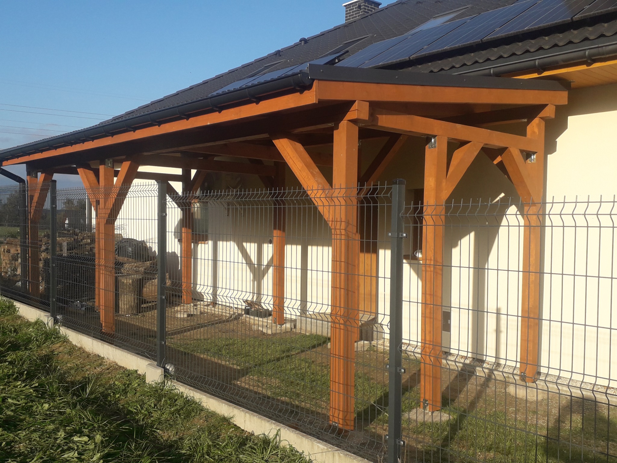 Drewniana konstrukcja zadaszenia tarasu przy domu jednorodzinnym, widoczna od strony ogrodu, z panelami słonecznymi na dachu budynku.