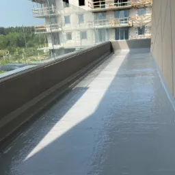 Świeżo wylana, szara, hydroizolacyjna powłoka na balkonie z szklaną balustradą, w tle budynek w stanie surowym otwartym.